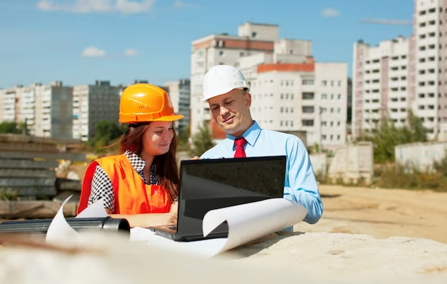 Two individuals wearing safety helmets and vests, viewing a laptop outdoors.